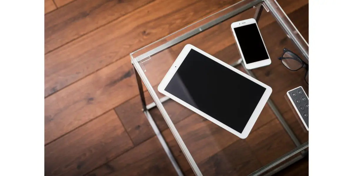 AdobeStock_123680240 iphone ipad next to remote and glasses on glass table on dark wood flooring