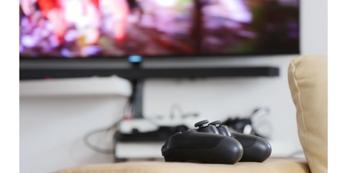 AdobeStock_133895463 ps4 controller on couch arm rest with ps4 game, tv blurred background