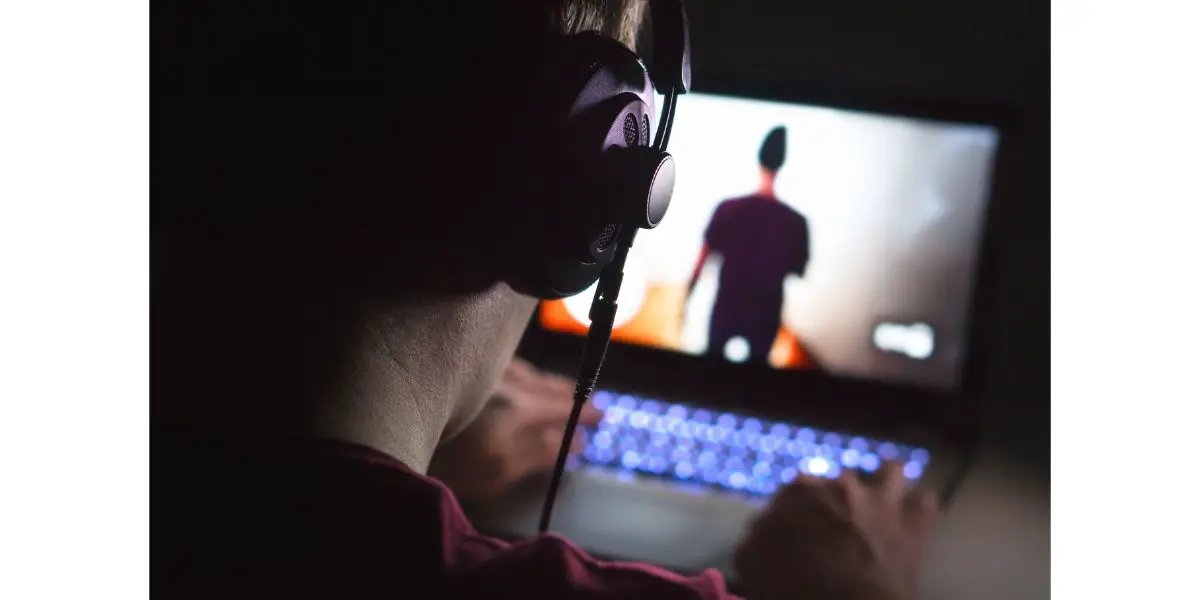 AdobeStock_191746050 Playing video games with laptop. Young man plays action game on computer. Back view of gamer with headphones in dark or late at night