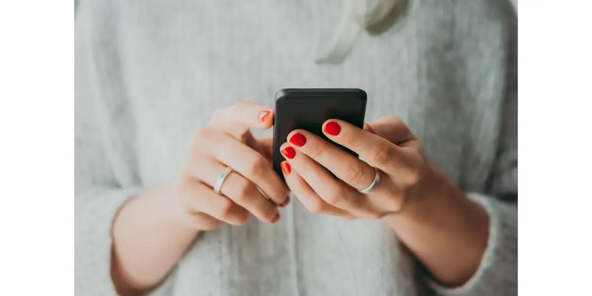 AdobeStock_228091226 woman with painted red nails, wearing 1 ring on each hang holding a black smartphone while wearing a grey sweater