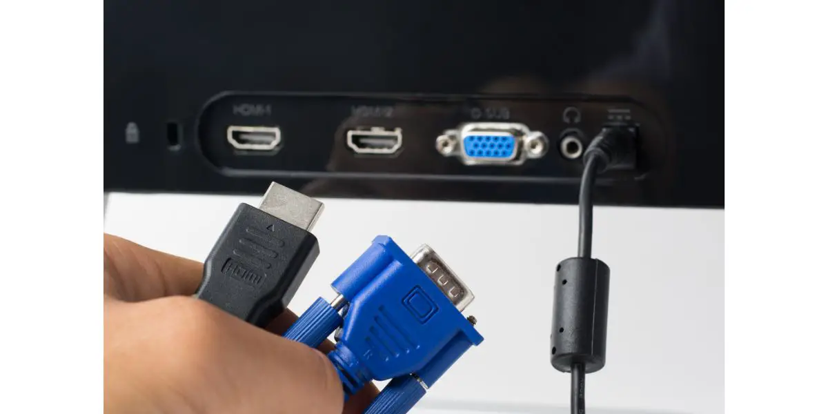 AdobeStock_243516462 Man`s hand holds HDMI and VGA cables against a monitor with ports. Choise between modern HDMI and old VGA connection