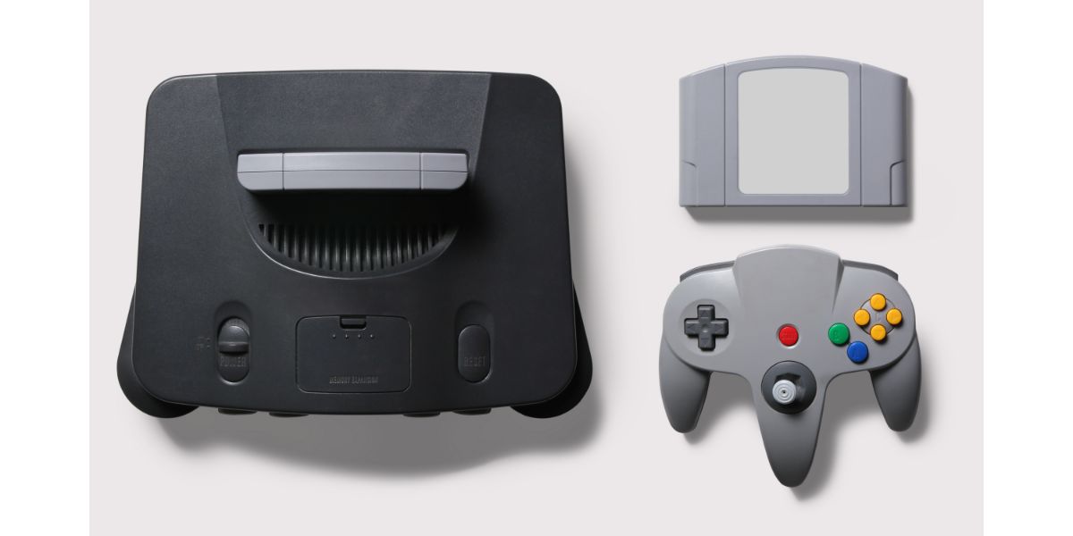 AdobeStock_371189778 Black N64 gaming consoler with grey controller joypad and game on a white background