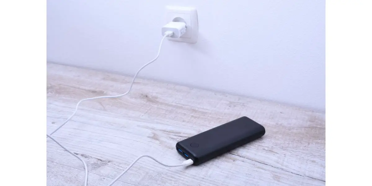 AdobeStock_395527151 Black phone power bank plugged in the socket on the wall for charging on wooden background