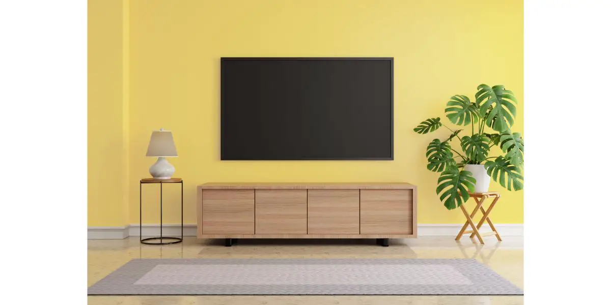 AdobeStock_425845729 Living room with blank screen hanging LCD Television mock up on yellow wall. Monstera plant and desk lamp and grey carpet on marble floor.