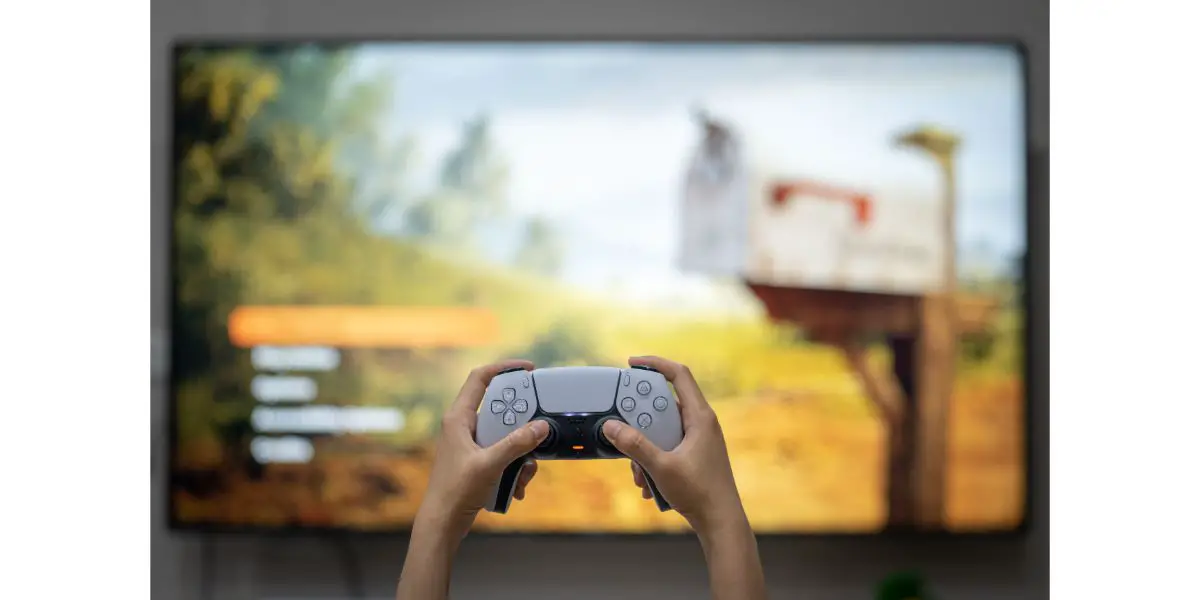 AdobeStock_429560321_Editorial_Use_Only Hands holding Play Station 5 controller with blurred background of It Takes Two game displayed on large tv