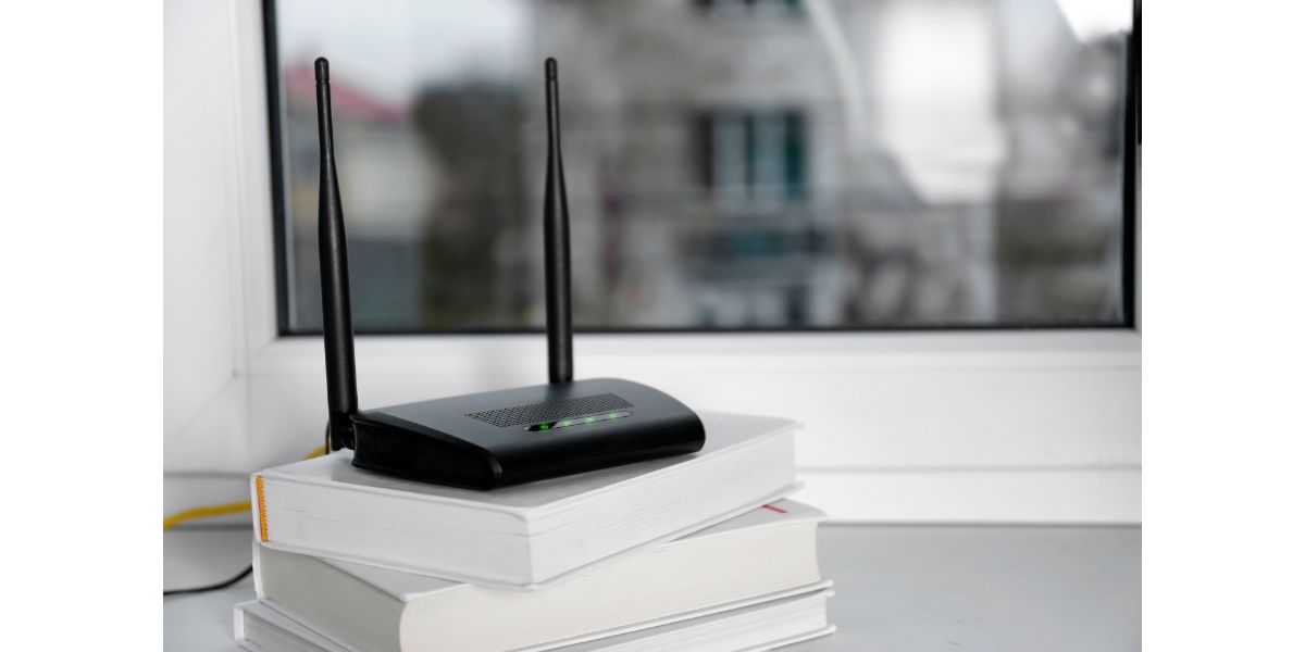 Modern Wi-Fi router on stack of books near window indoors.