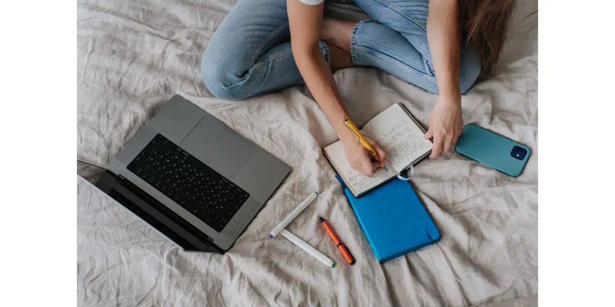AdobeStock_578022027 Cropped view of girl writing in diary sitting on bed with laptop phone pen, remote learning home. Top view on remote working young woman planning agenda at bedroom. 