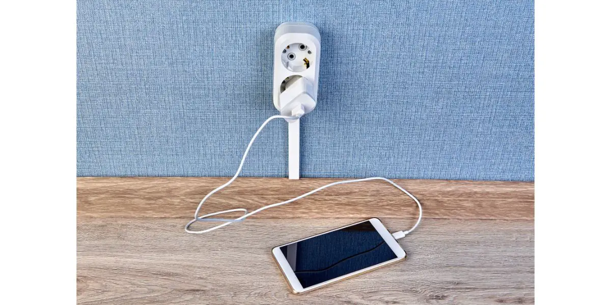 Depositphotos_257155800_L Mobile phone is plugged in socket on wood floow next to blue wall