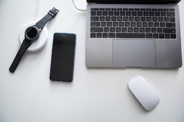 Depositphotos_354852348_S Laptop with phone computer mouse and smart watch charging on wireless charger on white table